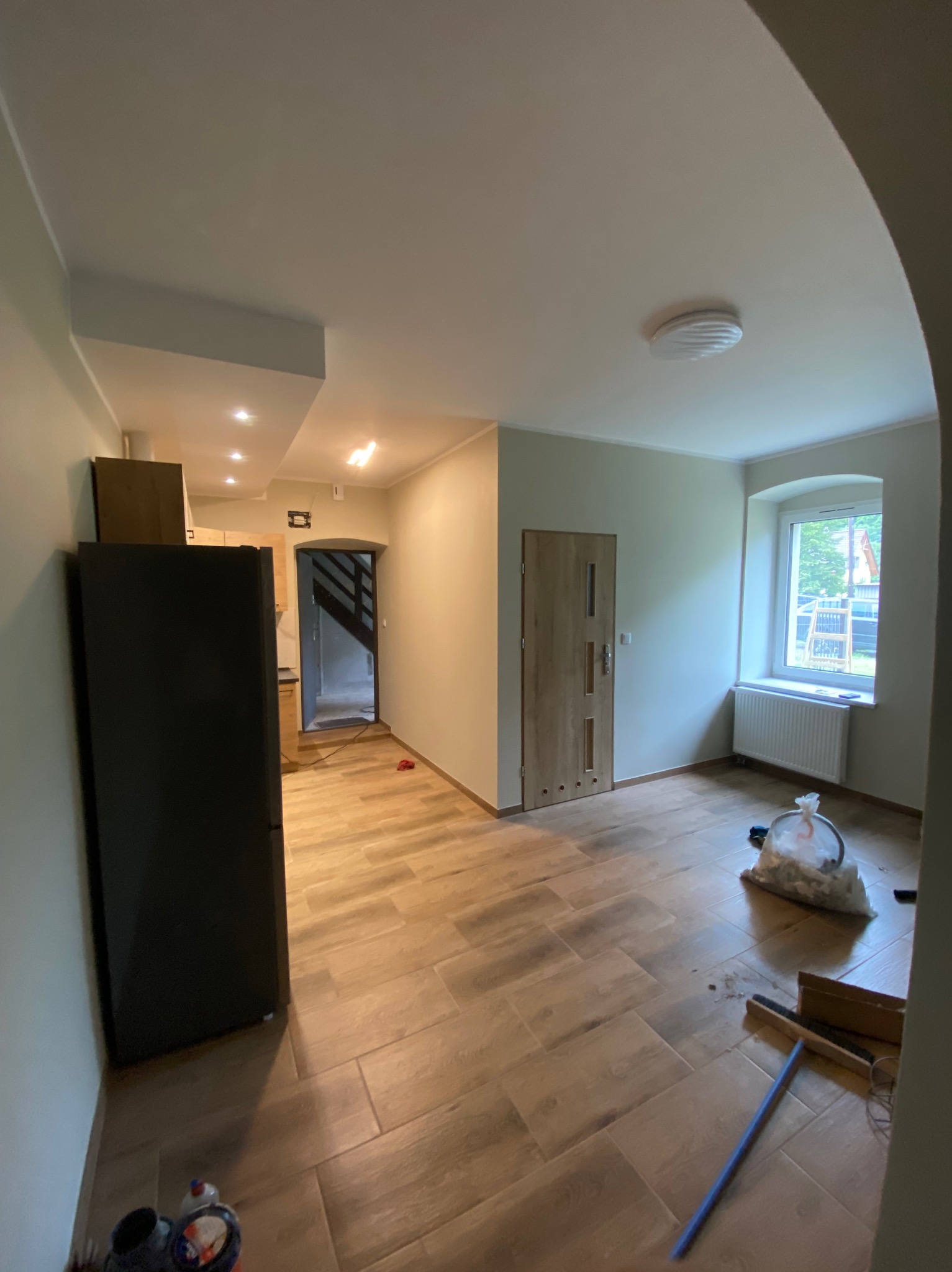 Wnętrze po remoncie: brązowe panele podłogowe ułożone w jodełkę, świeżo pomalowane ściany w odcieniach bieli, okno z grzejnikiem i widokiem na zieleń. Częściowo widoczna lodówka.