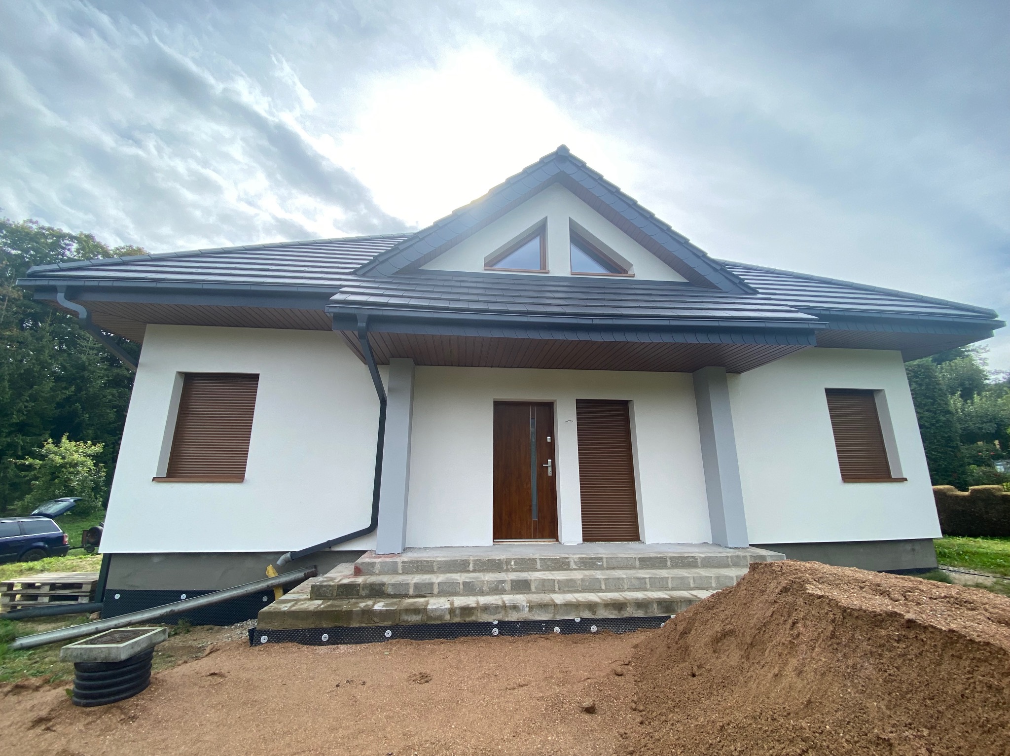 Nowoczesny dom jednorodzinny z białymi ścianami, brązowymi roletami i ciemną dachówką, widok z przodu na schody wejściowe i kopiec ziemi.