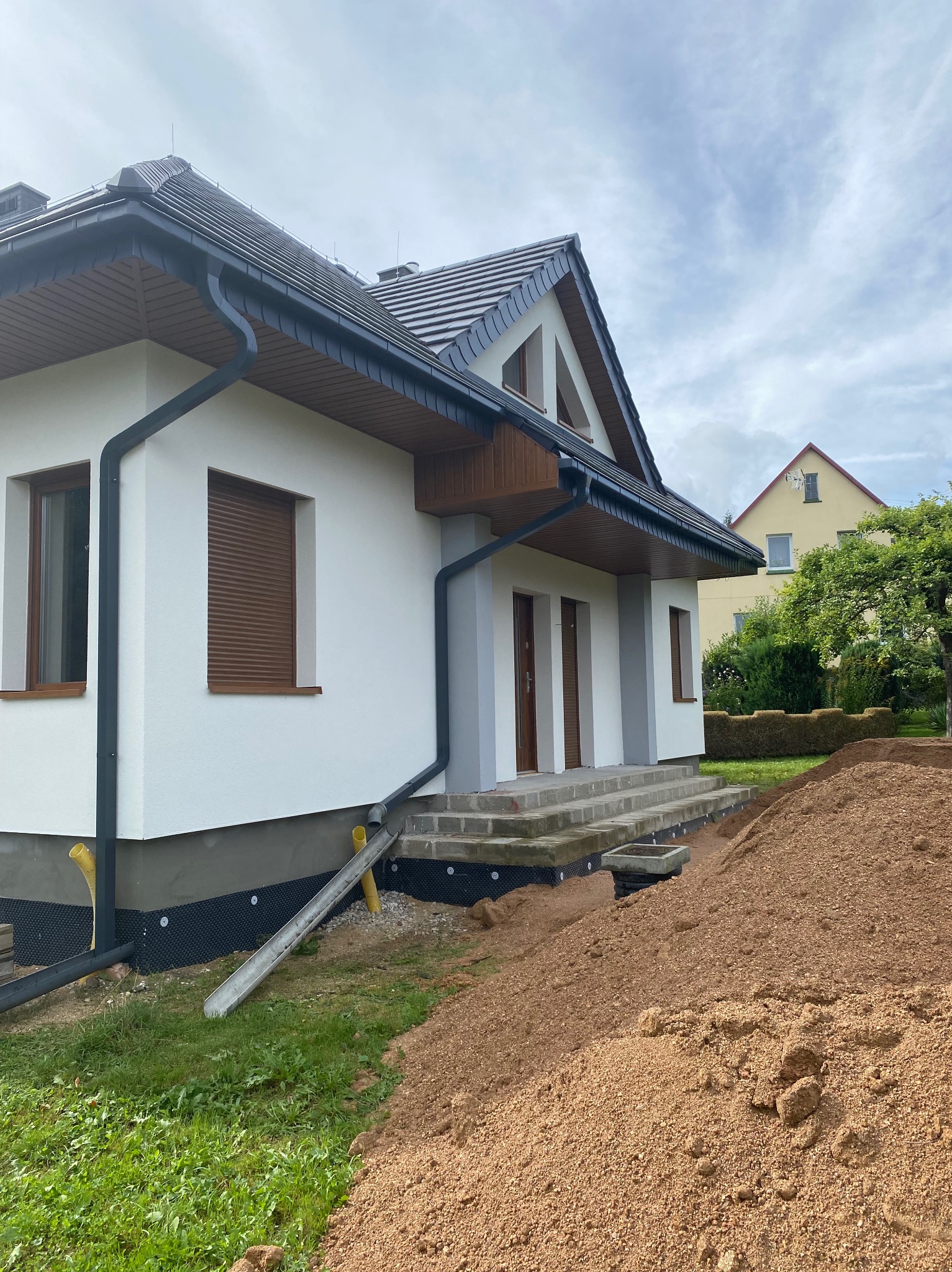 Biały dom jednorodzinny z szarą dachówką i brązowymi oknami, widok z boku na elewację i przygotowany teren pod ogród. Nowe orynnowanie.