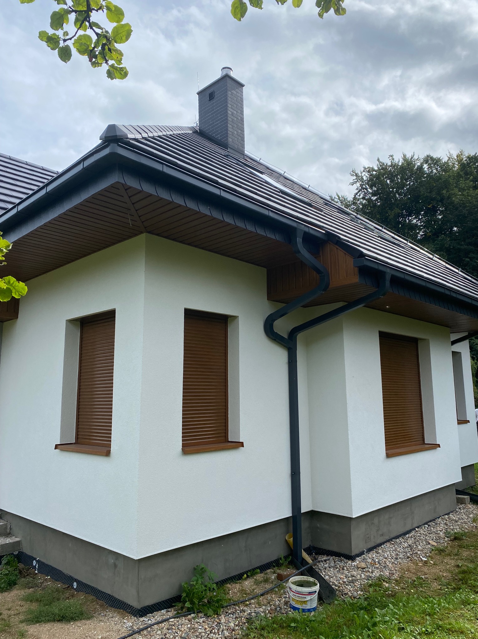 Boczny widok domu po renowacji elewacji, z brązowymi roletami okiennymi i ciemną dachówką, na tle pochmurnego nieba i zieleni.