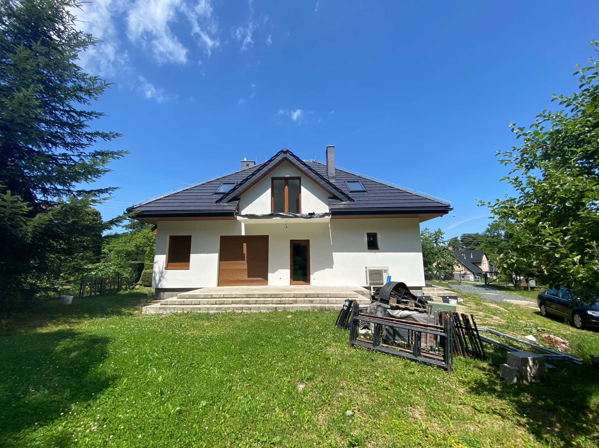 Biały dom jednorodzinny z ciemną dachówką i brązowymi okiennicami, otoczony zielonym trawnikiem i drzewami, w słoneczny dzień. Na trawniku leżą elementy konstrukcyjne.