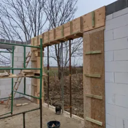 Wznoszenie ścian z bloczków betonowych, widoczne szalunki drewniane nad otworem drzwiowym, rusztowanie i wiadro na budowie w pochmurny dzień.