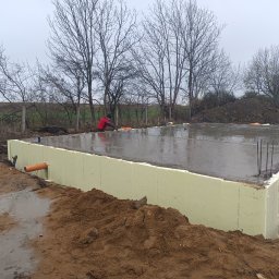 TJ Usługi Budowlane - Wylany fundament z izolacją termiczną z polistyrenu ekstrudowanego, wystającymi prętami zbrojeniowymi i pomarańczową rurą drenażową, widok na plac budowy w pochmurny dzień.