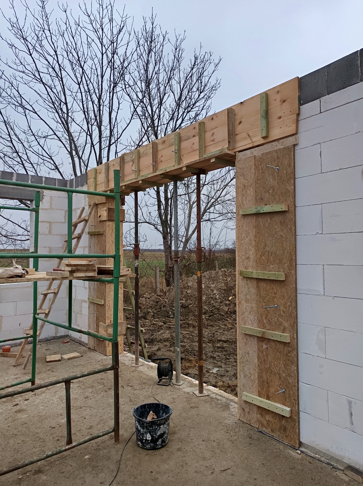 Wznoszenie ścian z bloczków betonowych, widoczne szalunki drewniane nad otworem drzwiowym, rusztowanie i wiadro na budowie w pochmurny dzień.