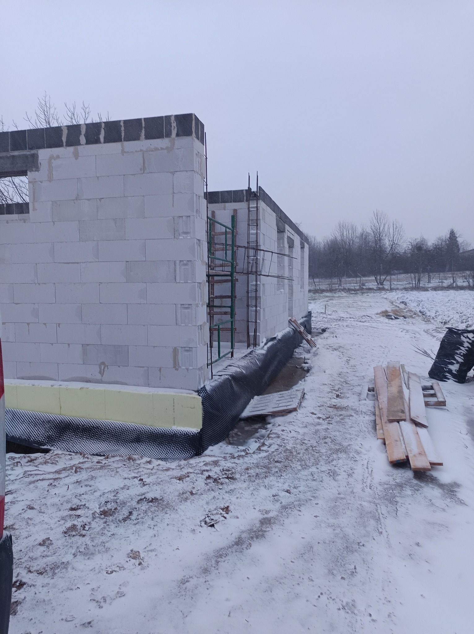 Surowy stan budynku z białych bloczków, widoczne rusztowanie, ocieplenie fundamentów i zasypany śniegiem teren budowy w pochmurny dzień.