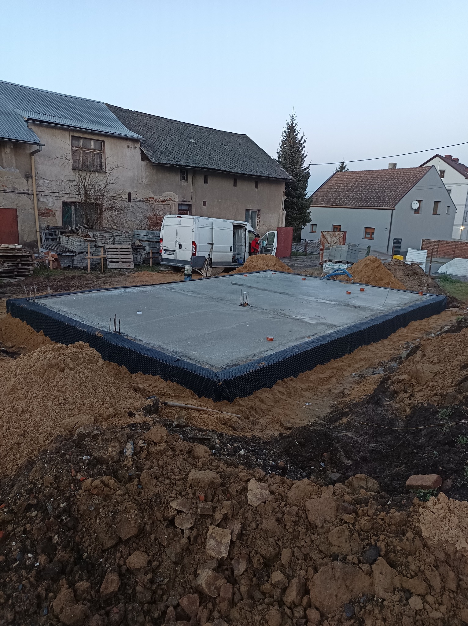 Zalany betonowy fundament z hydroizolacją w wykopie, widoczne wystające pręty zbrojeniowe i okoliczne zabudowania.