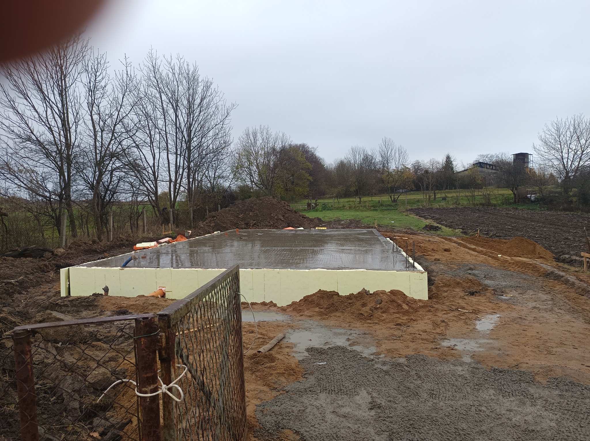 Zalany betonem fundament zabezpieczony styropianem, widok zza metalowej siatki ogrodzeniowej, w tle jesienny krajobraz.