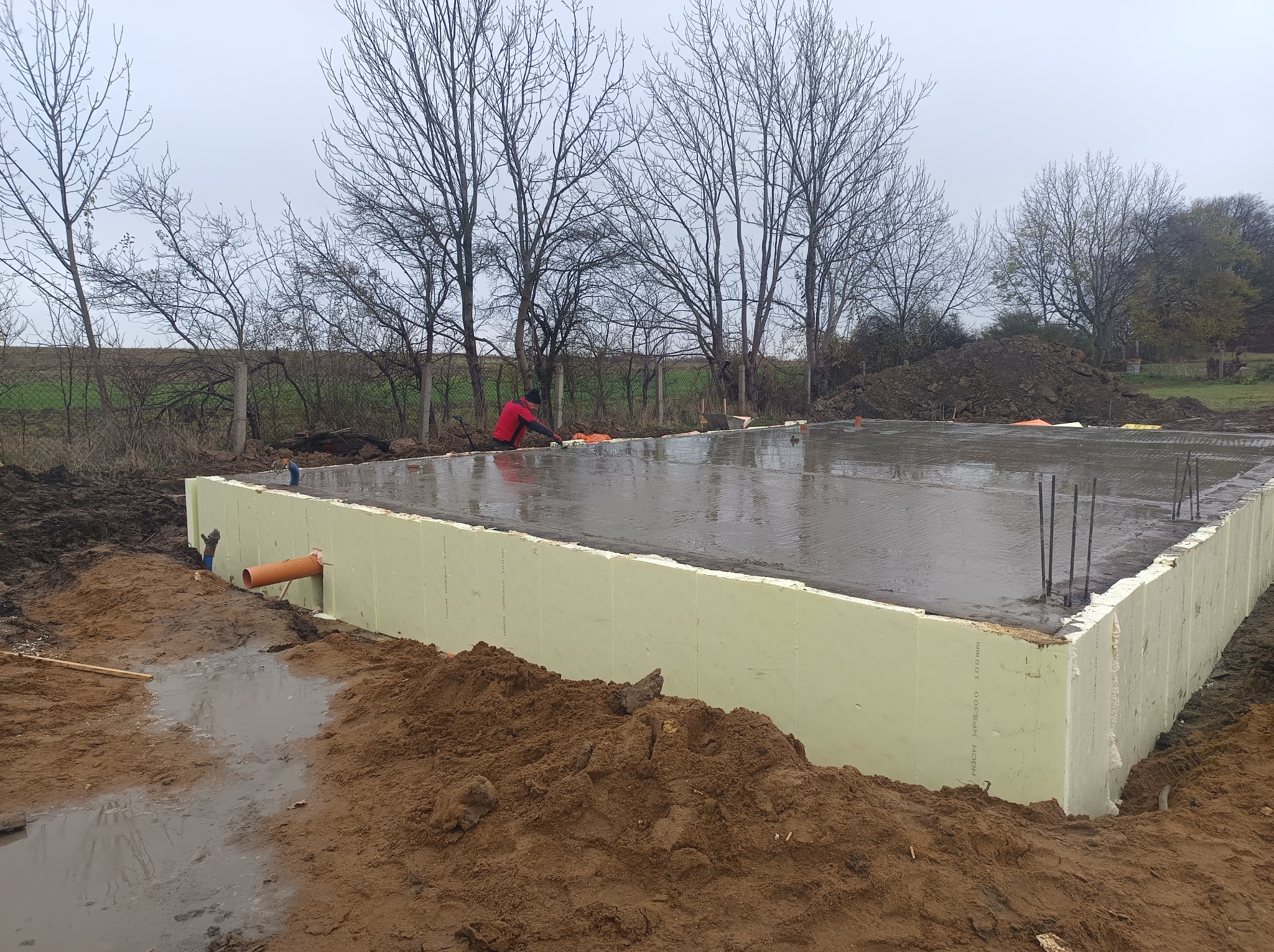 Wylany fundament z izolacją termiczną z polistyrenu ekstrudowanego, wystającymi prętami zbrojeniowymi i pomarańczową rurą drenażową, widok na plac budowy w pochmurny dzień.