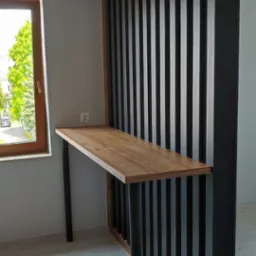 Drewniany blat biurka wsparty na czarnych, metalowych nogach, przymocowany do dekoracyjnej ścianki działowej z pionowych, czarnych listew na drewnianej ramie, widok z boku na tle okna.