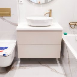 MILAR PROJEKT Waldemar Kruszelnicki - Nowoczesna łazienka z białą umywalką nablatową, beżową szafką pod umywalkę z dwoma szufladami, złotą armaturą i okrągłym lustrem z widocznym grzejnikiem drabinkowym w odbiciu.