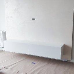 MILAR PROJEKT Waldemar Kruszelnicki - Biała, minimalistyczna szafka RTV zawieszona na ścianie z dekoracyjnym tynkiem w jasnym, nowoczesnym salonie, podłoga zabezpieczona kartonem, widoczny grzejnik i fragment pufy z metalowymi nóżkami.