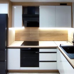 MILAR PROJEKT Waldemar Kruszelnicki - Nowoczesna kuchnia z białymi szafkami i drewnianym blatem, czarna lodówka Haier po lewej stronie, czarny zlew i bateria przy oknie z folią ochronną.