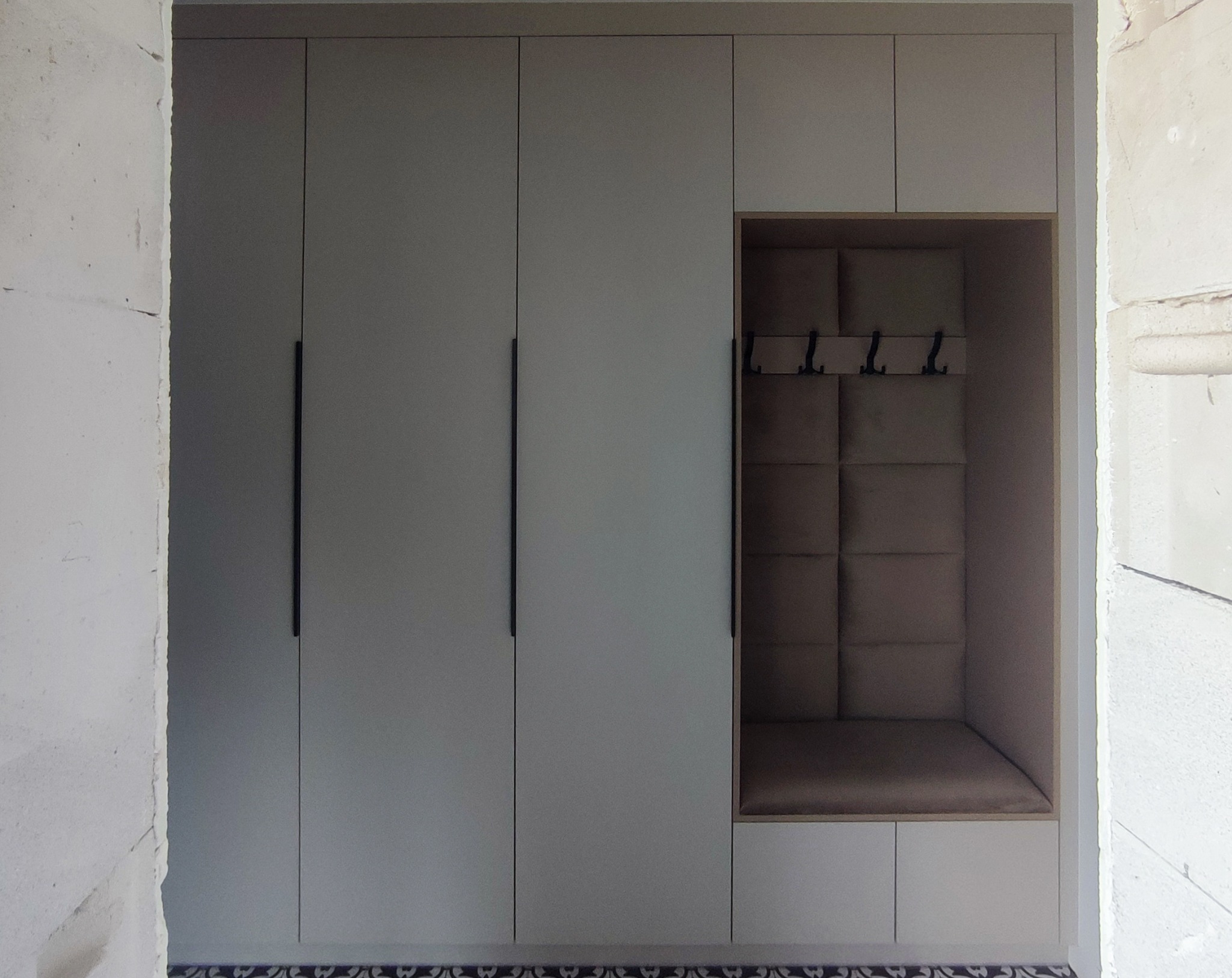 Szara zabudowa meblowa z szafami i wnęką z wieszakami i tapicerowanym panelem ściennym, widok na surową ścianę z boku.