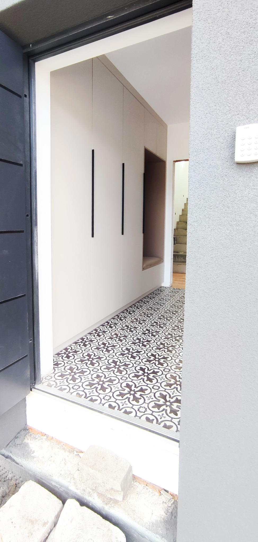 Wnętrze przedpokoju z zabudowaną szafą wnękową z kremowymi frontami i długimi, czarnymi uchwytami, wnęka z siedziskiem, podłoga wyłożona wzorzystymi płytkami.