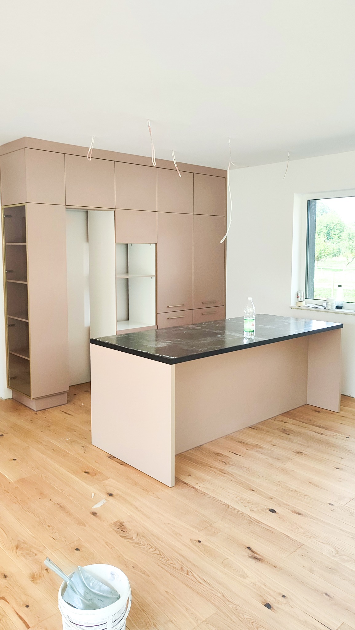 Montaż mebli kuchennych w jasnym pomieszczeniu: wysoka zabudowa w odcieniach beżu z otwartymi półkami i wyspa z czarnym blatem, widoczne kable zwisające z sufitu i wiadro z narzędziami na parkiecie.
