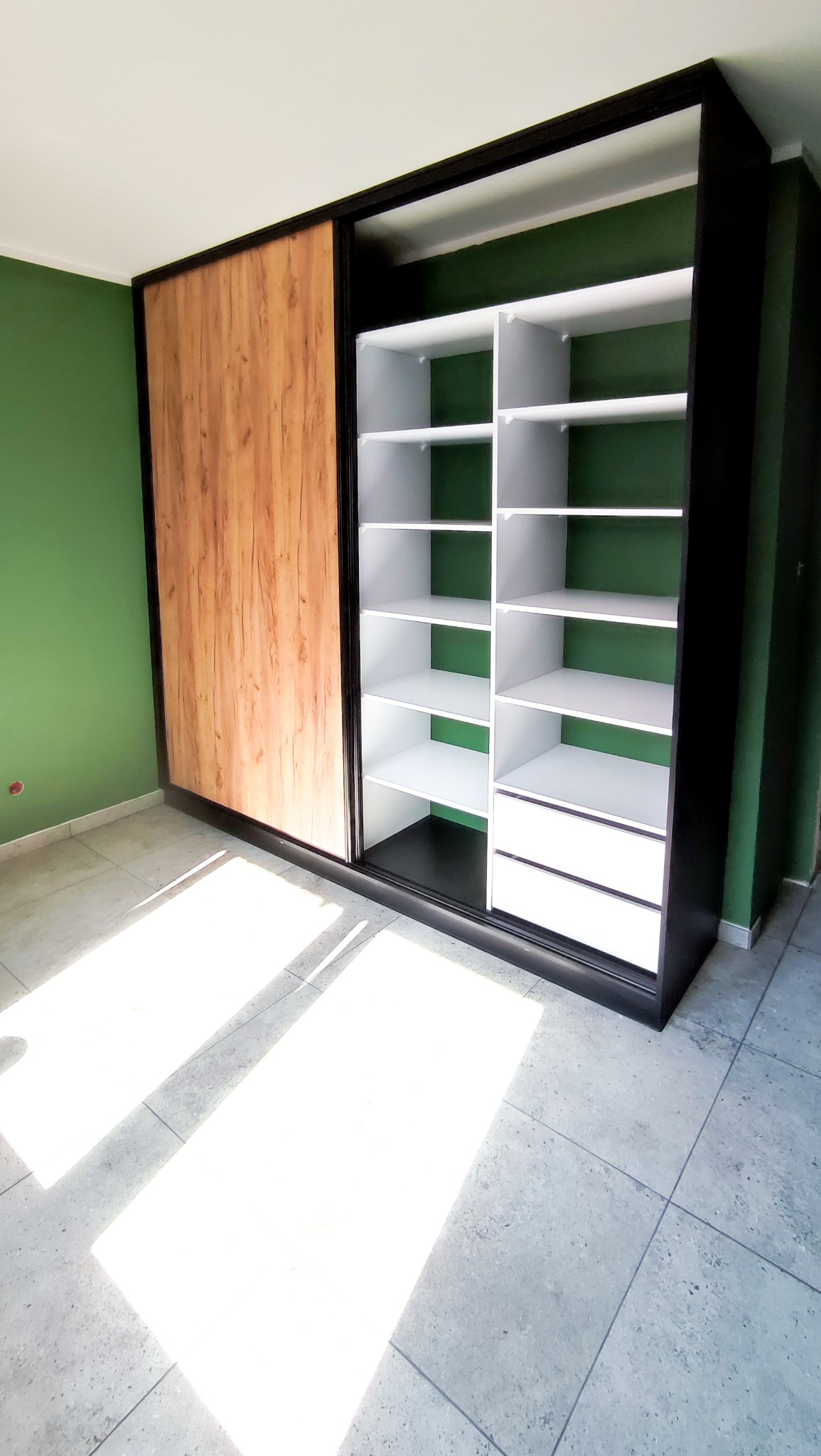 Wnętrze z szafą wnękową z przesuwnymi drzwiami w kolorze drewna i białymi półkami, czarna rama, zielona ściana w tle, jasne płytki na podłodze.