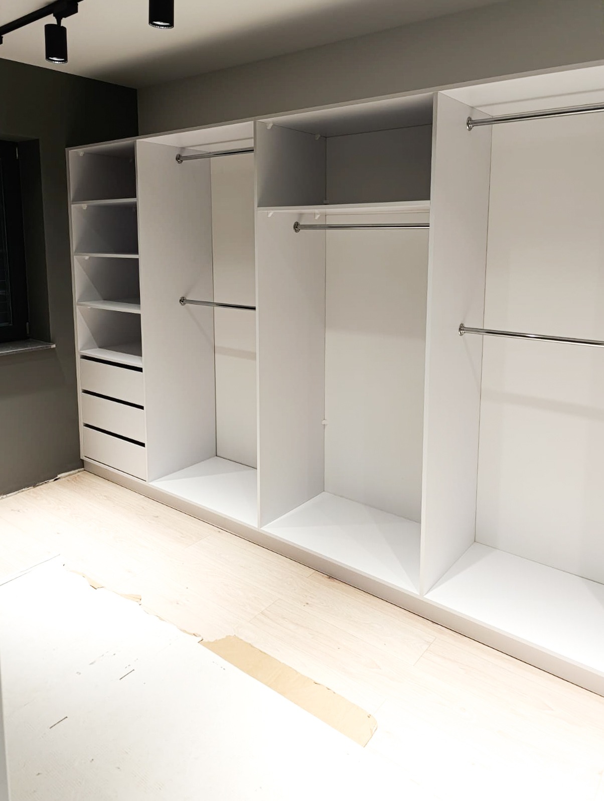 Biała szafa wnękowa z półkami i drążkami na ubrania, widoczne niedokończone panele podłogowe.