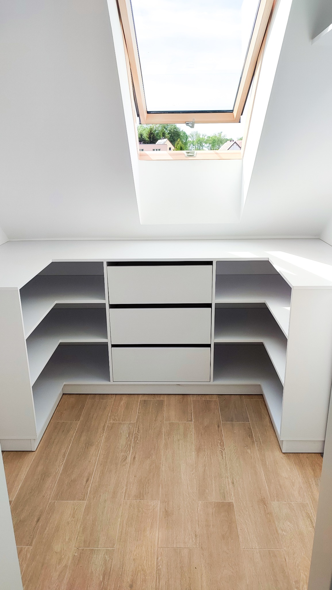 Biała szafa wnękowa z trzema szufladami i półkami, umieszczona na poddaszu z oknem dachowym, widok na drewnianą podłogę.