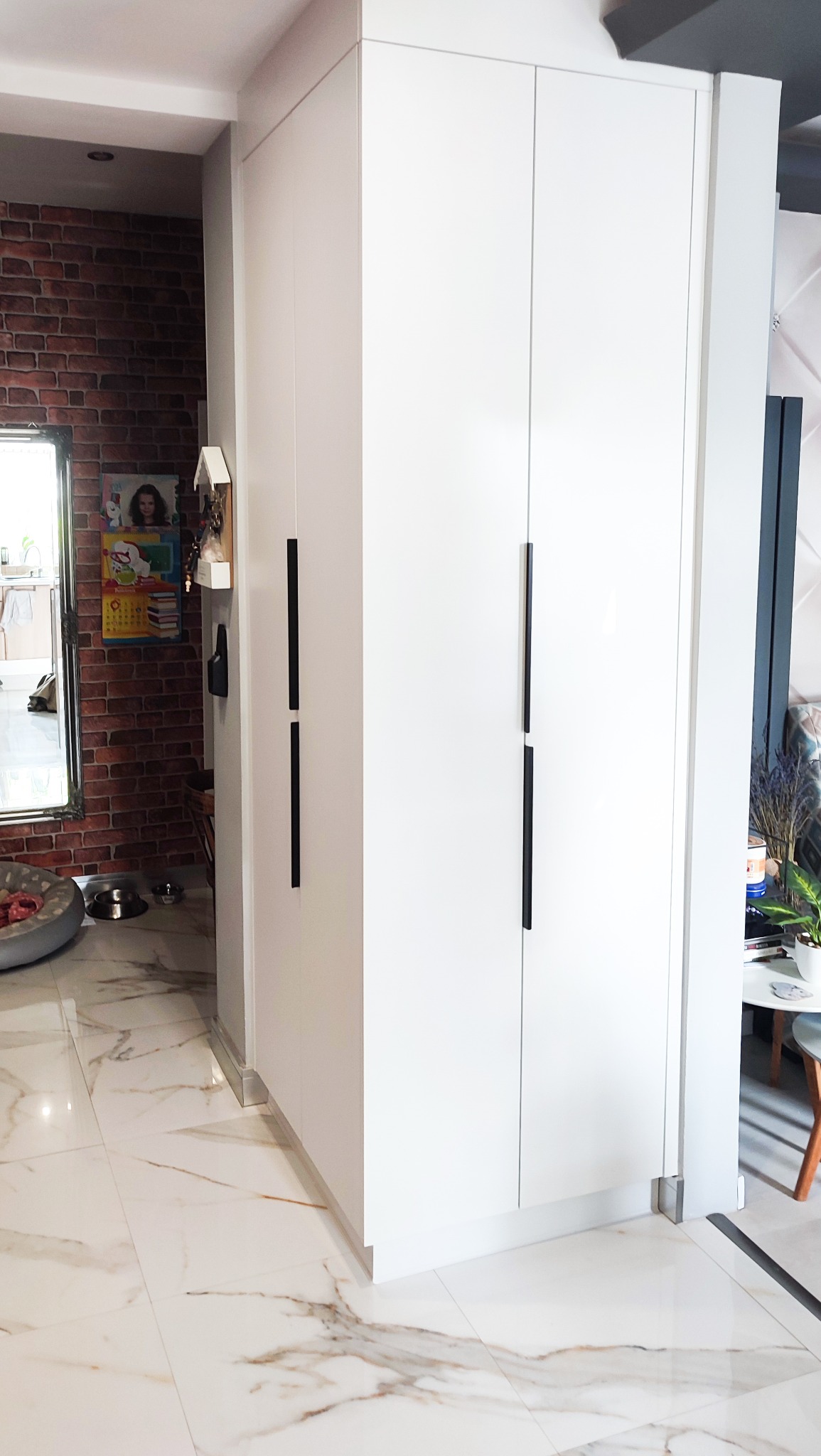 Biała szafa wnękowa z długimi, czarnymi uchwytami na tle marmurowej podłogi i ściany z czerwonej cegły w tle