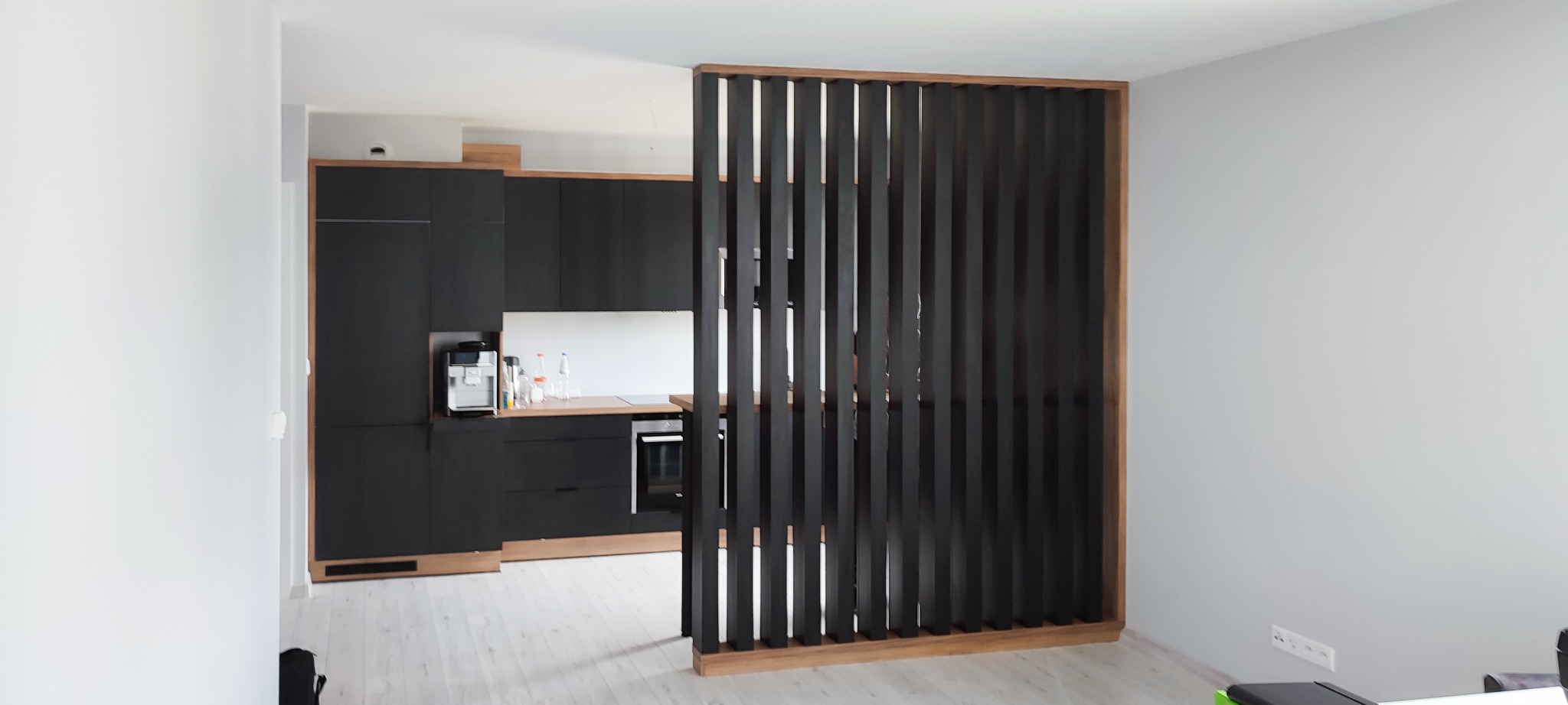 Nowoczesna kuchnia z czarnymi frontami i drewnianym blatem, oddzielona od reszty pomieszczenia ażurową, drewnianą ścianką działową z pionowych listew.