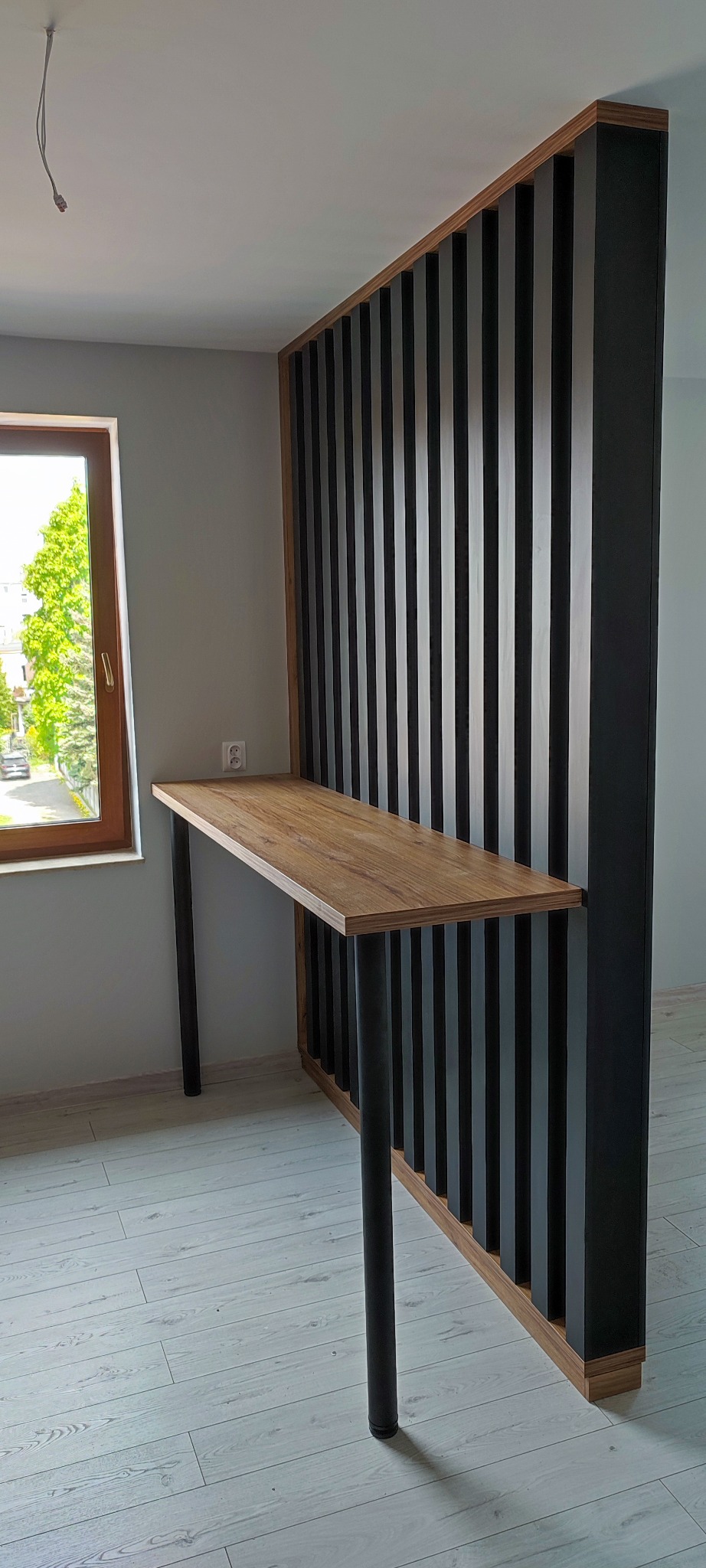Drewniany blat biurka wsparty na czarnych, metalowych nogach, przymocowany do dekoracyjnej ścianki działowej z pionowych, czarnych listew na drewnianej ramie, widok z boku na tle okna.