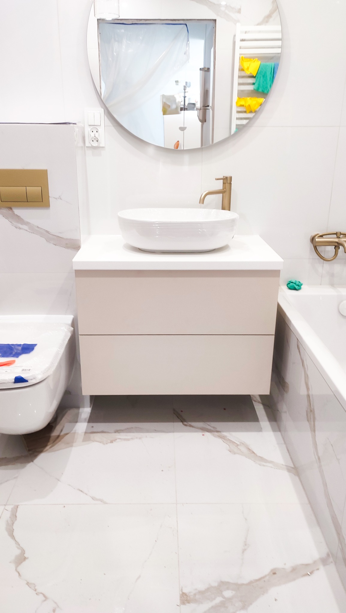 Nowoczesna łazienka z białą umywalką nablatową, beżową szafką pod umywalkę z dwoma szufladami, złotą armaturą i okrągłym lustrem z widocznym grzejnikiem drabinkowym w odbiciu.