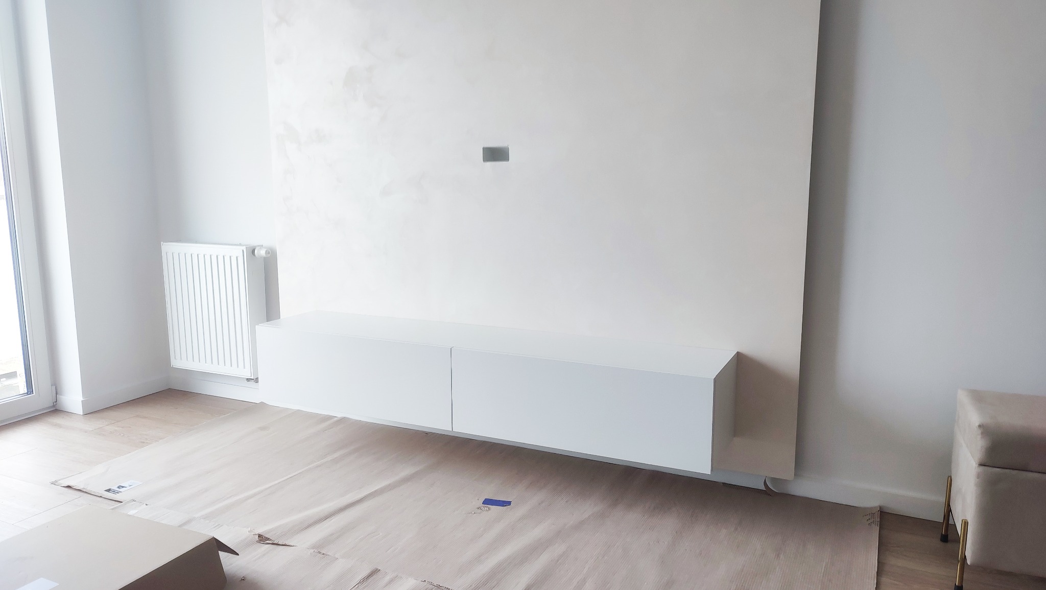 Biała, minimalistyczna szafka RTV zawieszona na ścianie z dekoracyjnym tynkiem w jasnym, nowoczesnym salonie, podłoga zabezpieczona kartonem, widoczny grzejnik i fragment pufy z metalowymi nóżkami.