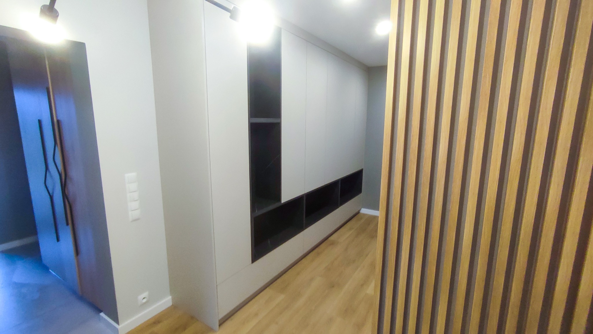 Nowoczesna zabudowa w przedpokoju: szara szafa z otwartymi półkami w dolnej i górnej części, drewniane lamele dekoracyjne na ścianie, drzwi z ciemnego drewna z podłużnymi uchwytami.