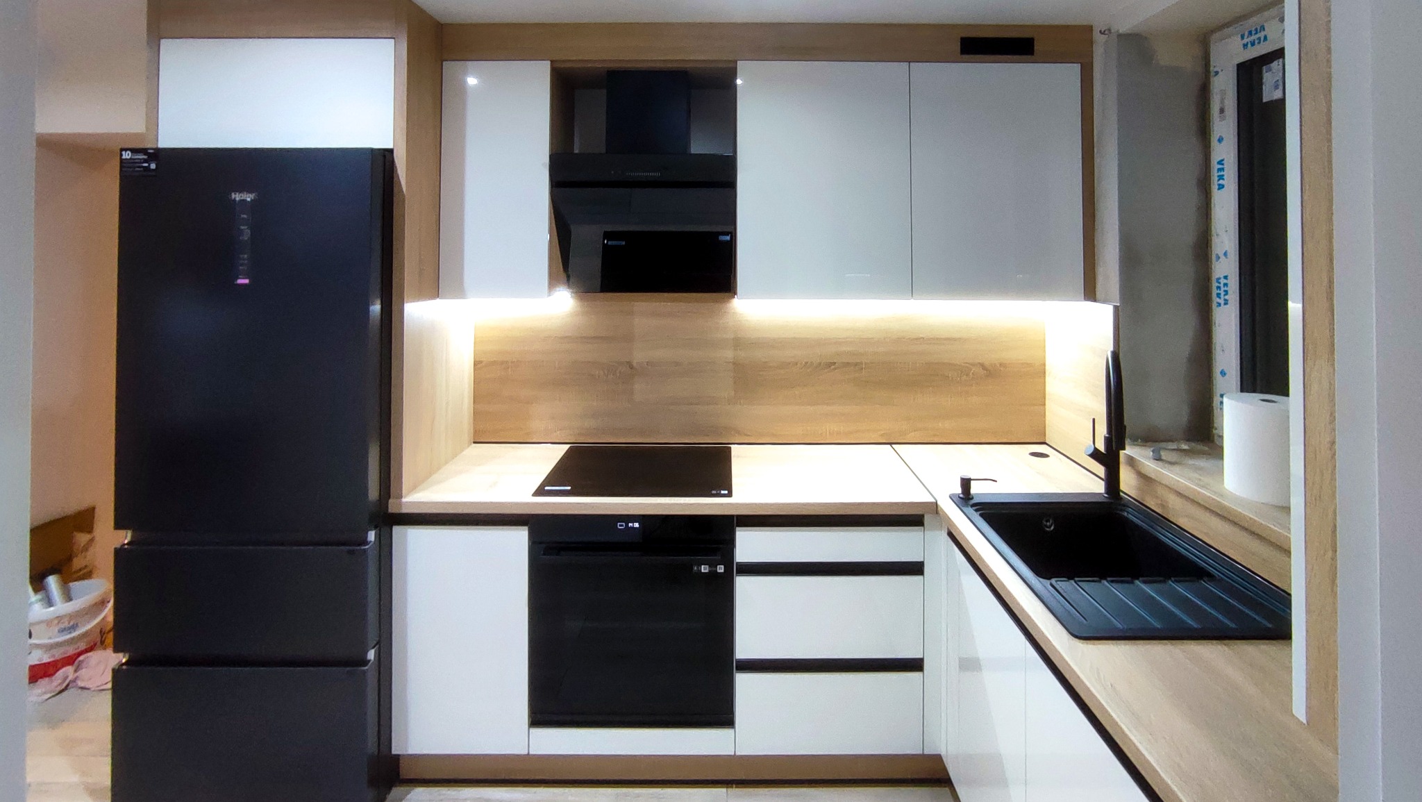 Nowoczesna kuchnia z białymi szafkami i drewnianym blatem, czarna lodówka Haier po lewej stronie, czarny zlew i bateria przy oknie z folią ochronną.