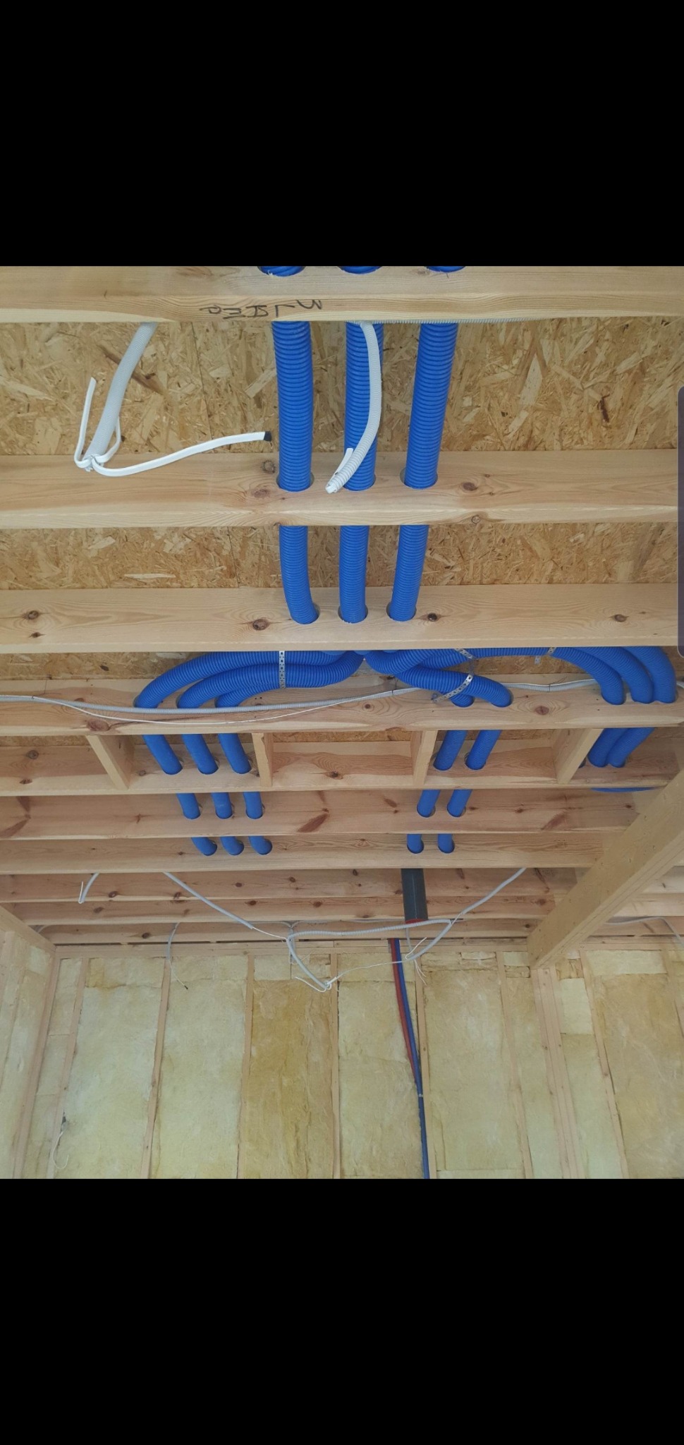 Niebieskie rury wentylacyjne, częściowo przymocowane metalowymi obejmami, prowadzone pod drewnianym stropem budynku w trakcie budowy; ściany ocieplone wełną mineralną.