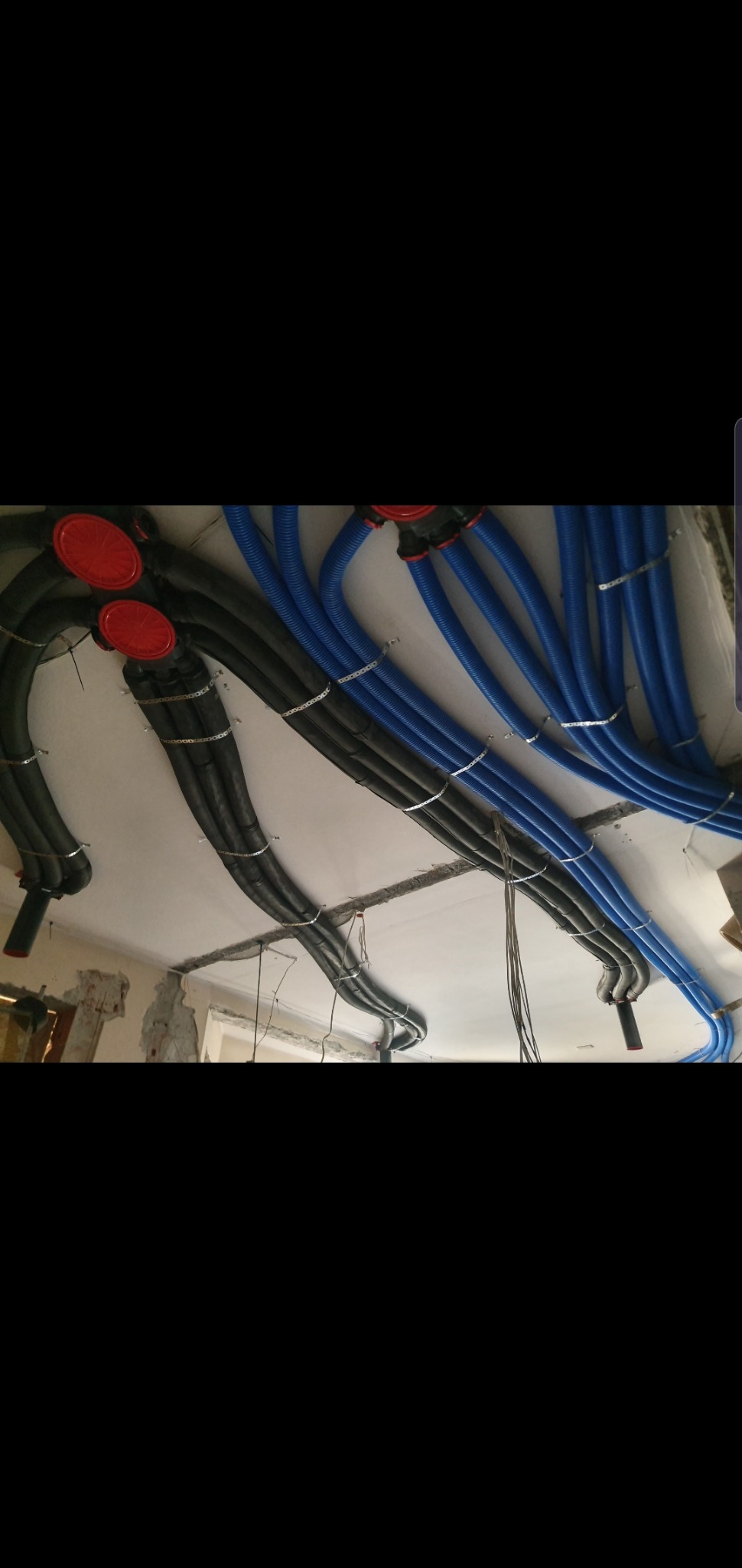 Niebieskie i czarne rury wentylacyjne przymocowane metalowymi obejmami do sufitu w trakcie montażu, widoczne czerwone elementy łączeniowe i fragment ściany w stanie surowym.