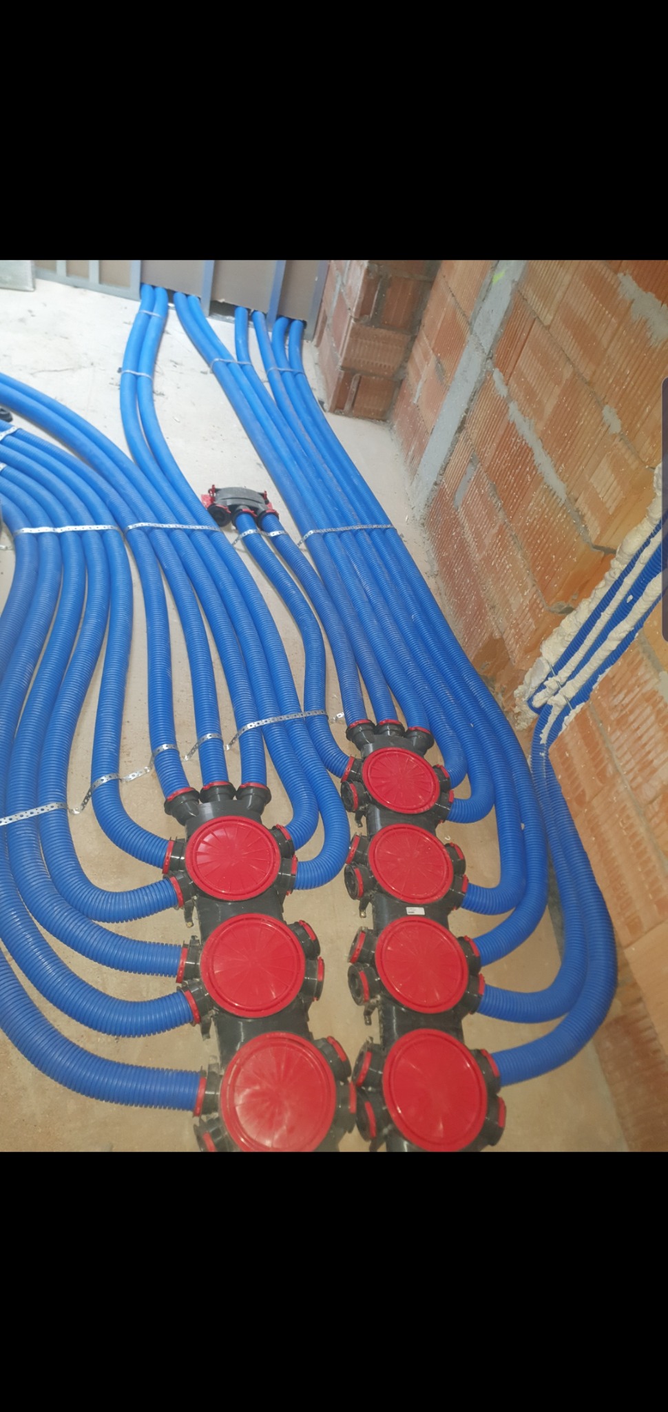 Szereg niebieskich, karbowanych rur wentylacyjnych podłączonych do czarnych rozdzielaczy z czerwonymi pokrywami, zamocowanych do ściany z czerwonej cegły.