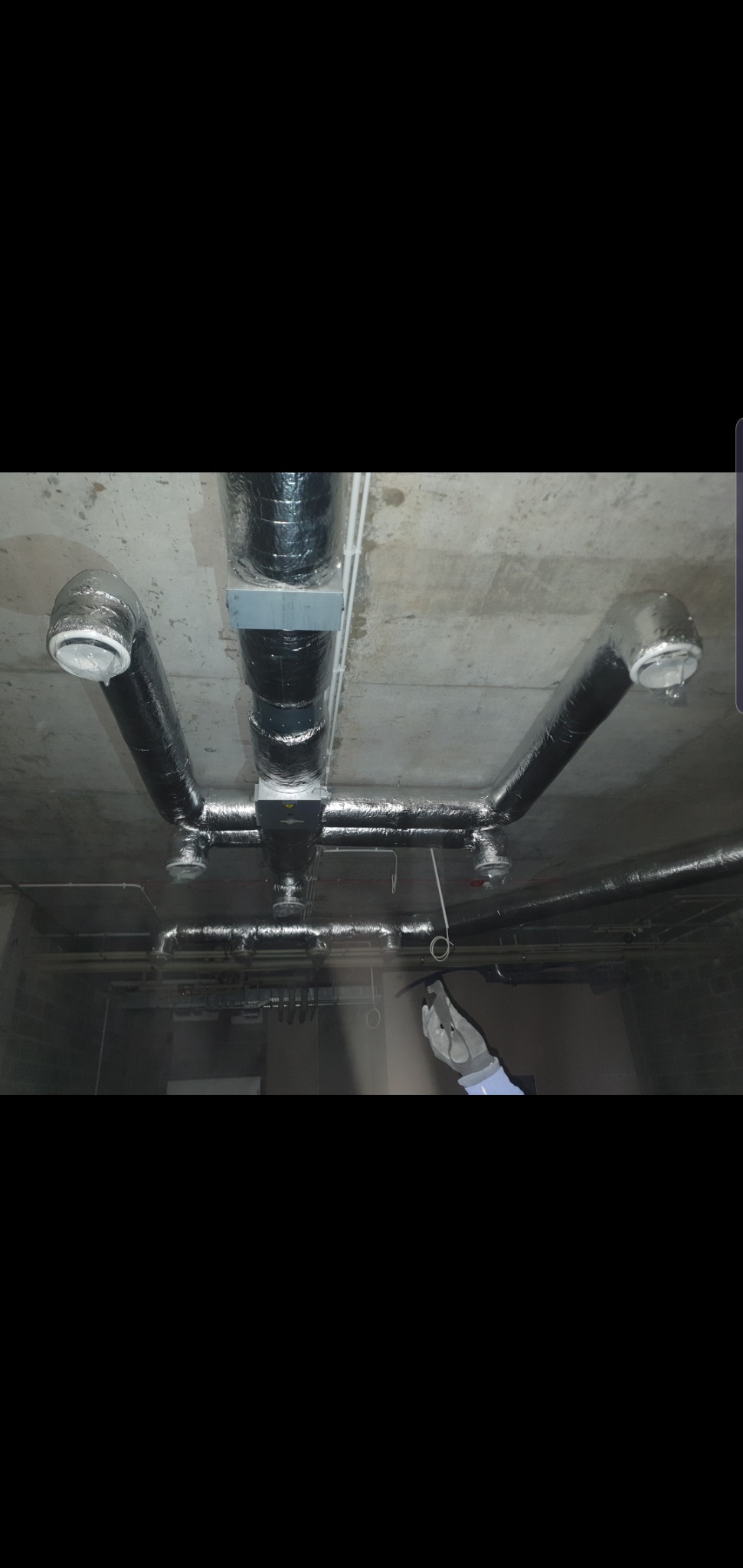 Instalacja przewodów wentylacyjnych w izolacji aluminiowej pod betonowym sufitem, widoczna ręka w roboczej rękawicy.