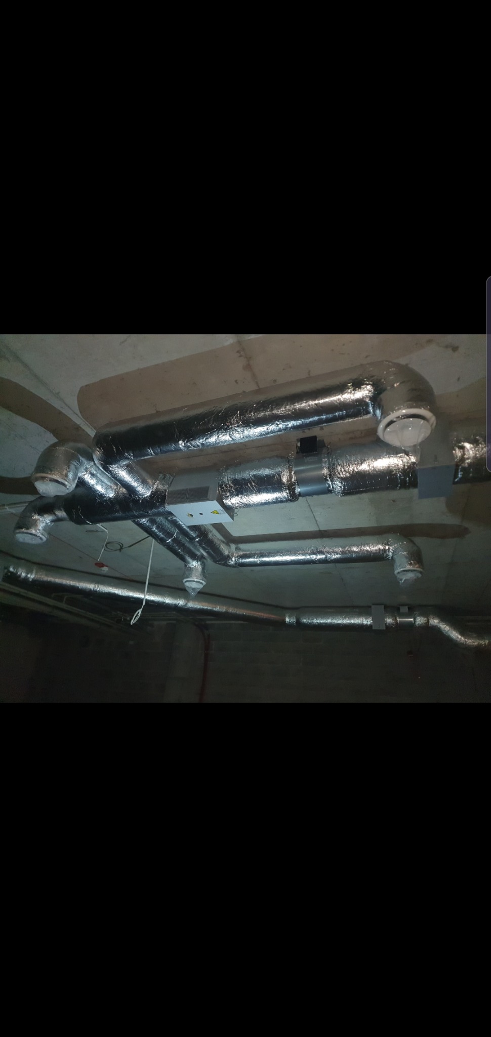 System rur wentylacyjnych i rekuperacyjnych, owiniętych srebrną folią izolacyjną, zamontowany pod betonowym sufitem w surowym stanie.