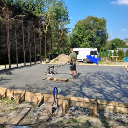 FHU JAN -KOP - Pracownik wygładza świeżo wylany beton na przygotowanym fundamencie za pomocą zacieraczki mechanicznej; drewniane szalunki widoczne wzdłuż krawędzi; w tle zaparkowany biały bus i drzewa.