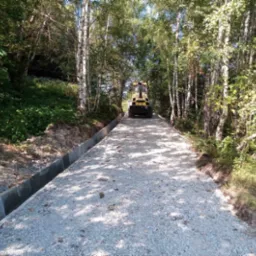 Żółty walec drogowy BOMAG ubija nowo wysypany żwir na drodze leśnej z betonowymi krawężnikami w słoneczny dzień.