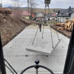 FHU JAN -KOP - Widok z kabiny maszyny budowlanej na układane betonowe płyty drogowe, teren przygotowany, w tle budynki mieszkalne i stosy drewna.