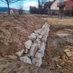 FHU JAN -KOP - Wznoszenie niskiego muru z szarych kamiennych bloków na nierównym, gliniastym terenie budowy, z widocznymi domami w tle i pochmurnym niebem.