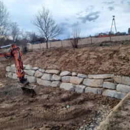 Pomarańczowa koparka układa warstwę z jasnych, nieregularnych kamieni na zboczu, tworząc mur, z widoczną ziemią i gruzem na pierwszym planie oraz drzewami i budynkami w tle pod pochmurnym niebem.