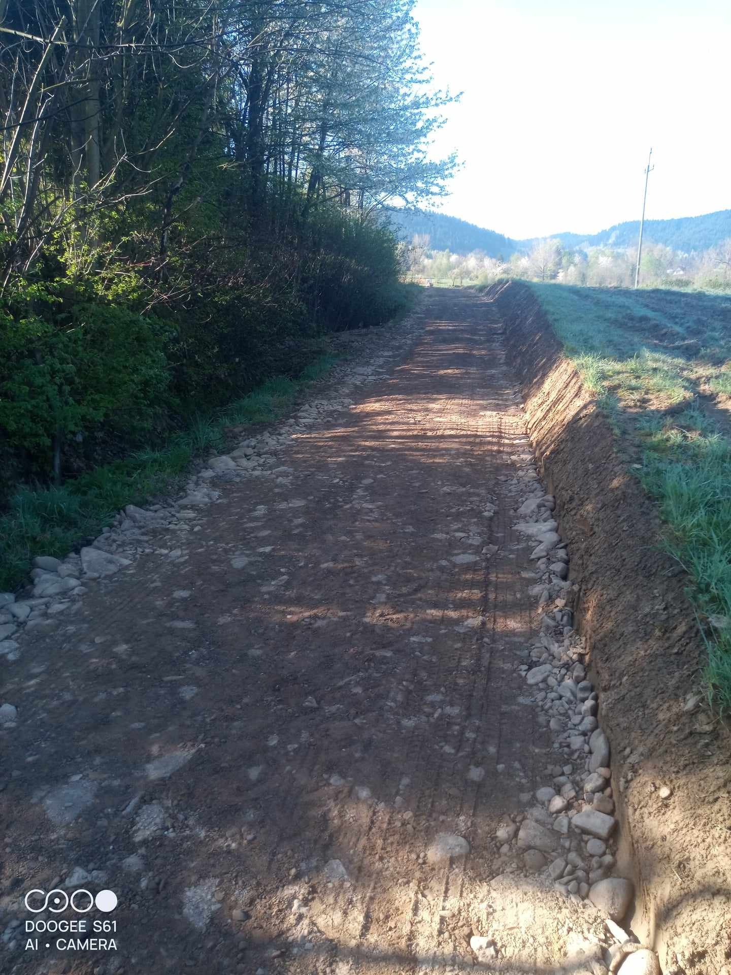 Wyrównany teren pod drogę gruntową z widocznymi śladami opon, obrzeżami z wykopanej ziemi i kamieniami po bokach, w tle zalesione wzgórza pod błękitnym niebem.