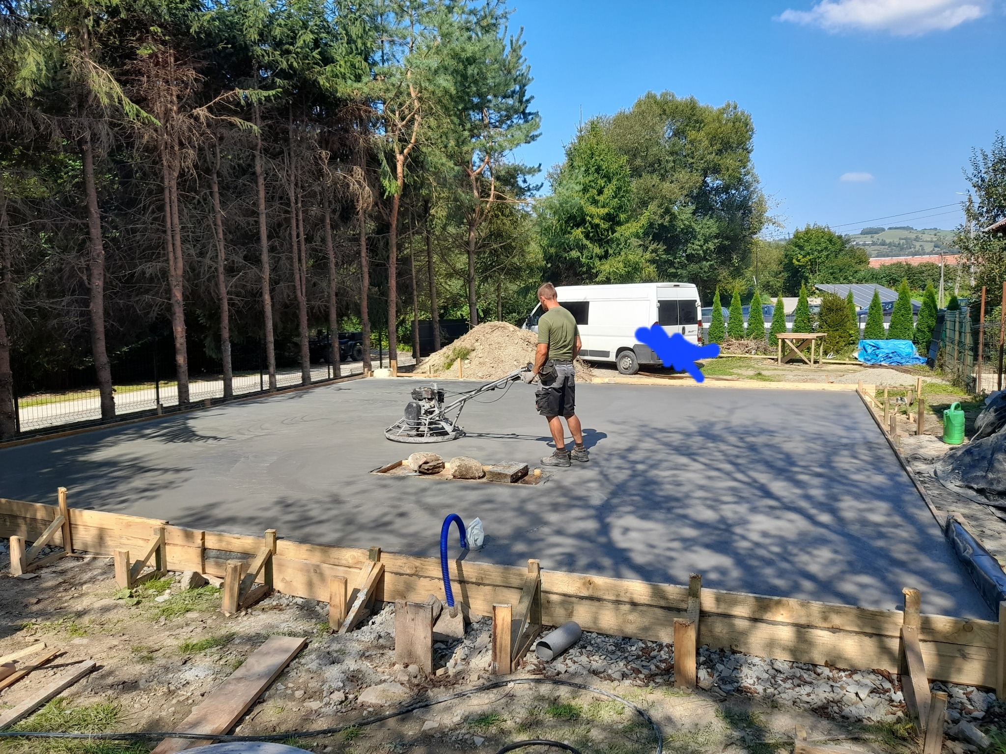 Pracownik wygładza świeżo wylany beton na przygotowanym fundamencie za pomocą zacieraczki mechanicznej; drewniane szalunki widoczne wzdłuż krawędzi; w tle zaparkowany biały bus i drzewa.