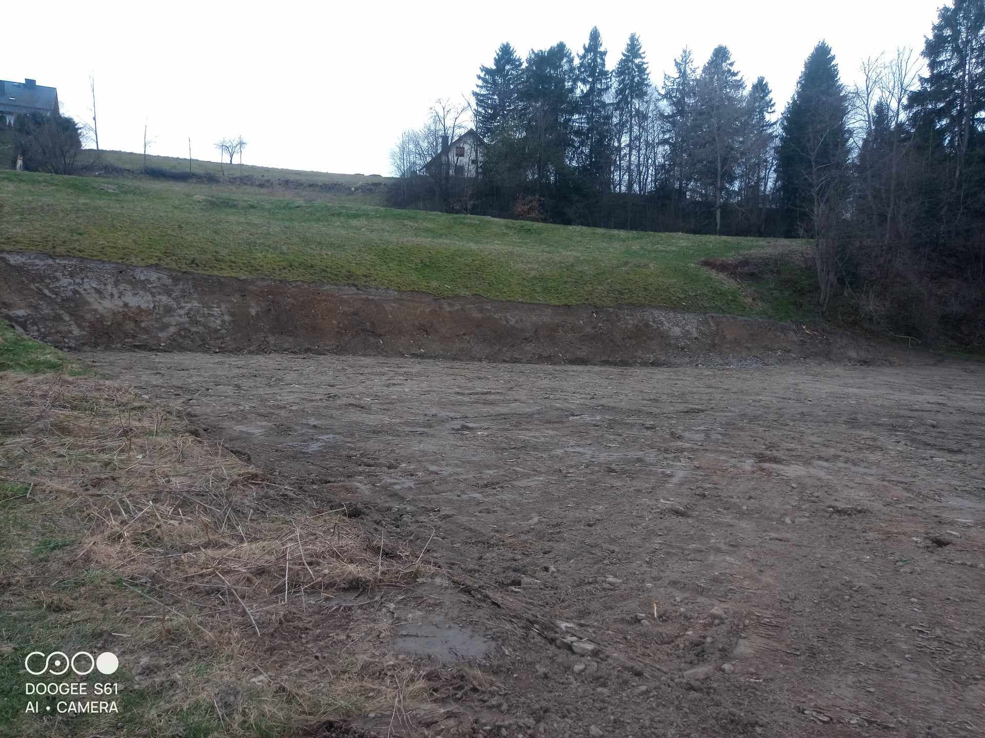 Przygotowany teren pod budowę z wykopem, widoczna warstwa humusu i odsłonięta gleba, w tle trawiasta skarpa i drzewa iglaste.