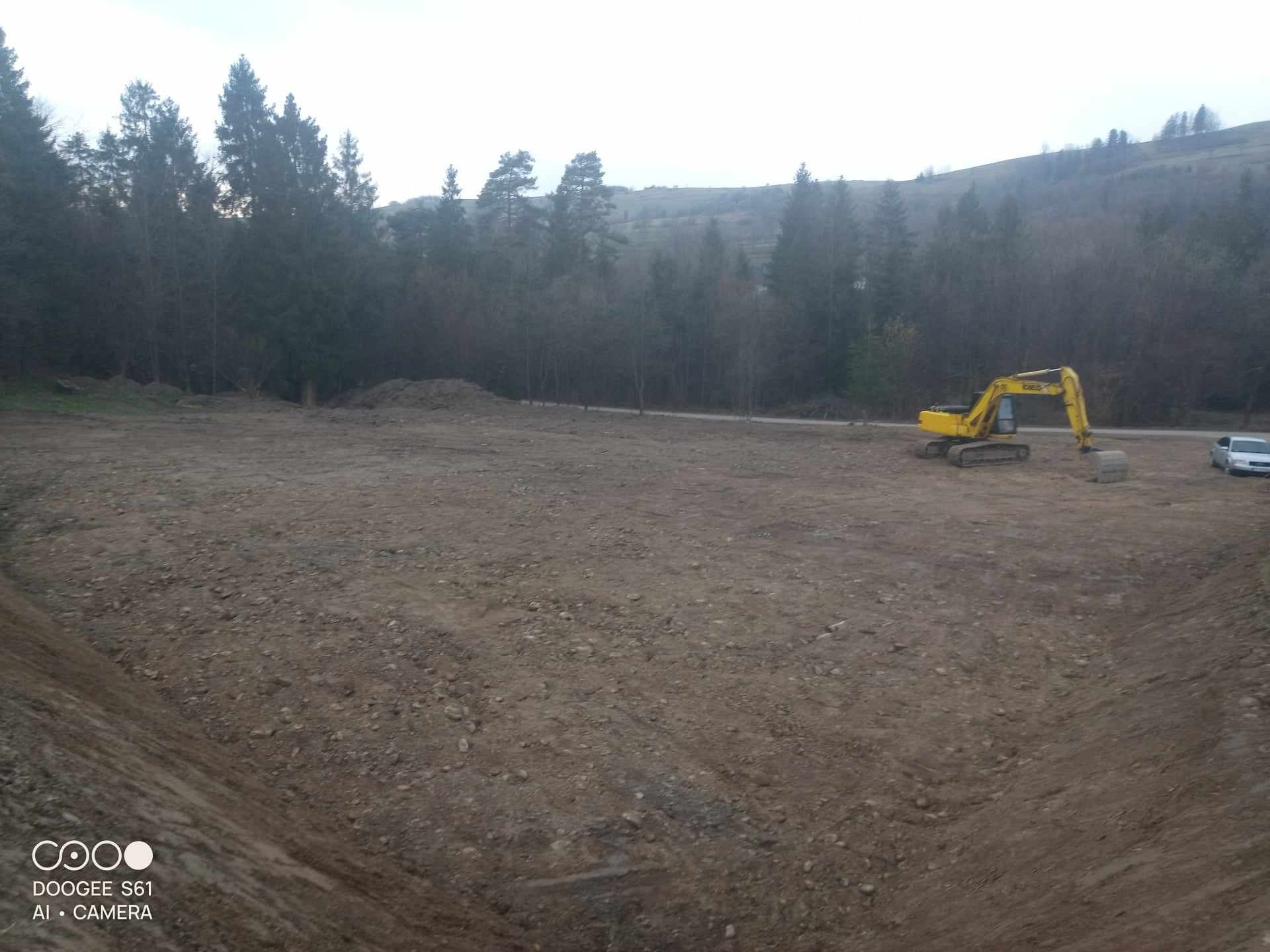 Żółta koparka gąsienicowa na rozległym, wyrównanym terenie z widocznym wykopem w tle lasu i wzgórz