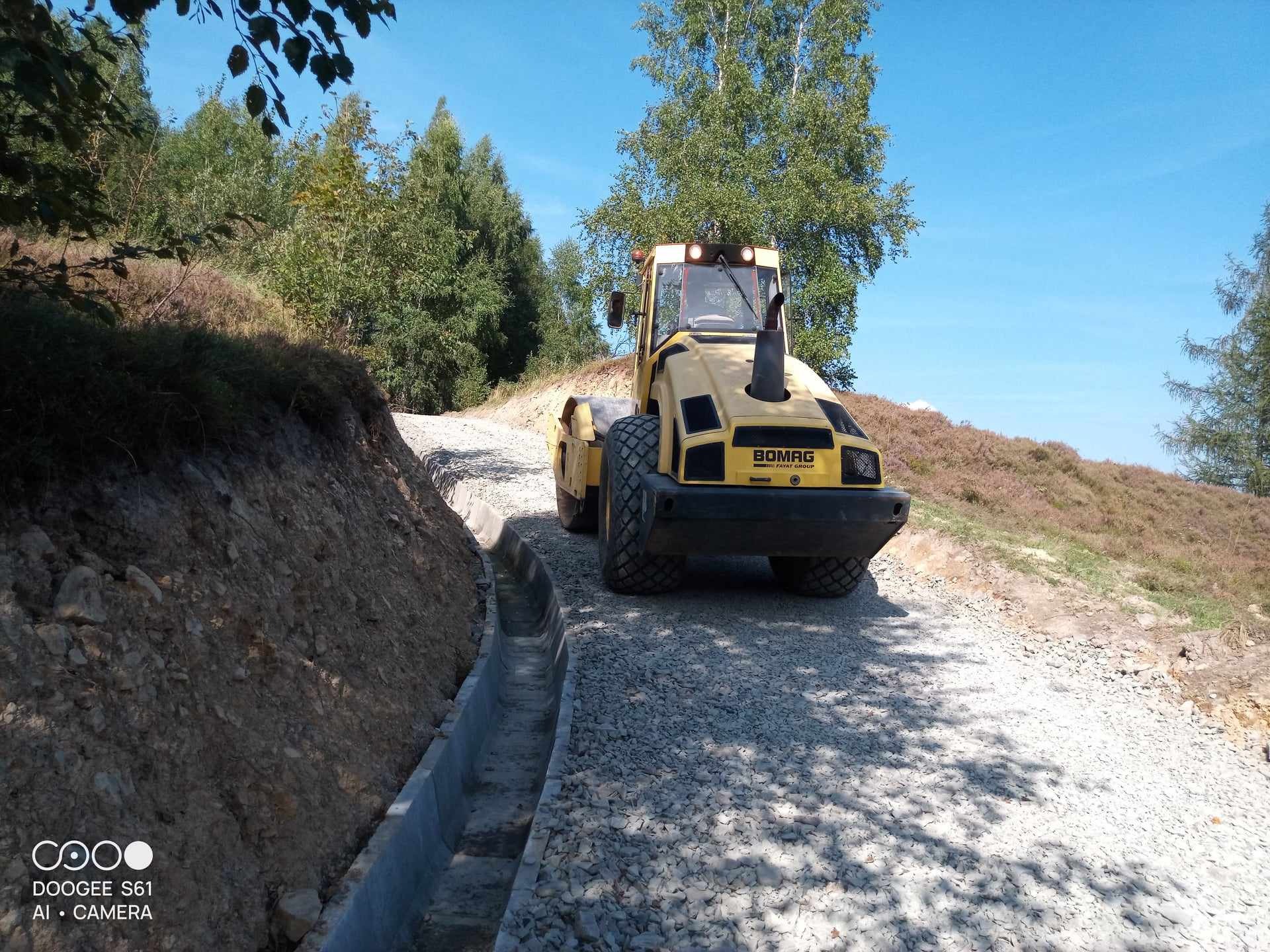 Żółty walec drogowy Bomag ubija szutrową drogę z betonowym korytkiem odwadniającym, w tle drzewa i pagórkowaty teren.