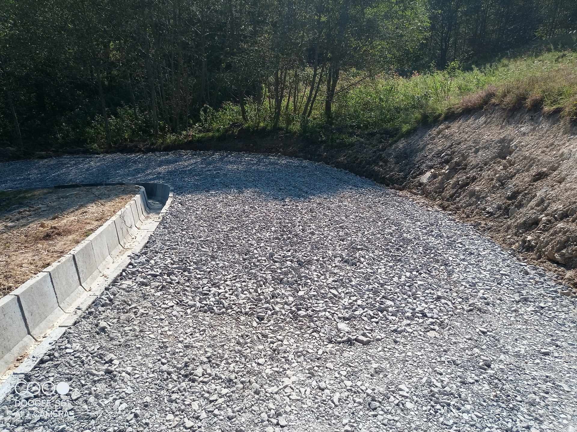 Świeżo utwardzona droga gruntowa z krawężnikiem betonowym i rowem odwadniającym, otoczona zielenią i skarpą ziemną.