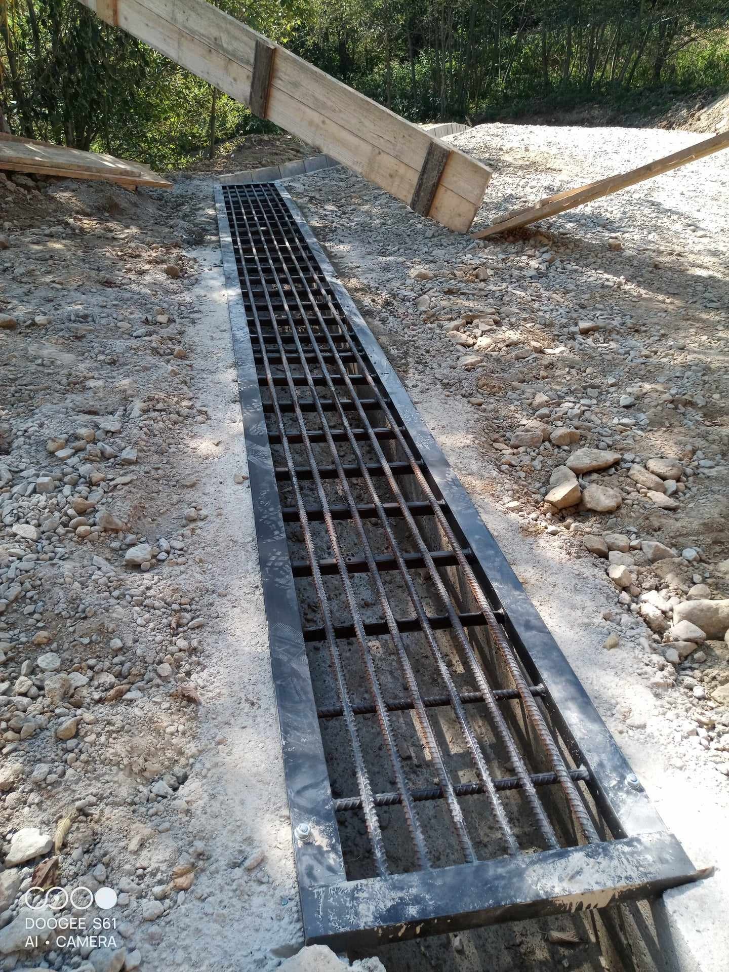 Długi, metalowy ruszt odwadniający w betonowej obudowie, ułożony w ziemi z kamieniami, z drewnianą konstrukcją wspierającą obok, zarośla w tle.
