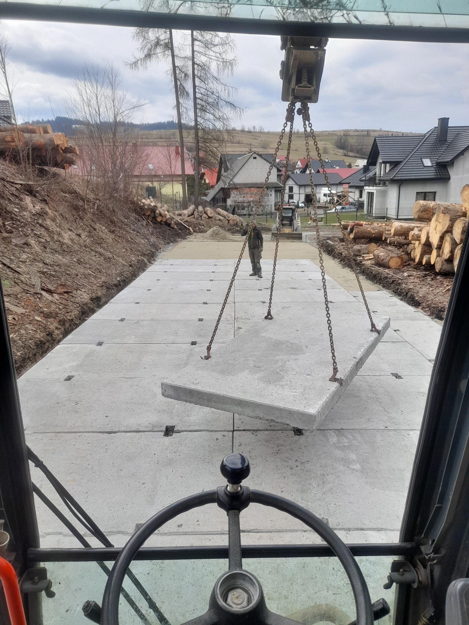 Widok z kabiny maszyny budowlanej na układane betonowe płyty drogowe, teren przygotowany, w tle budynki mieszkalne i stosy drewna.