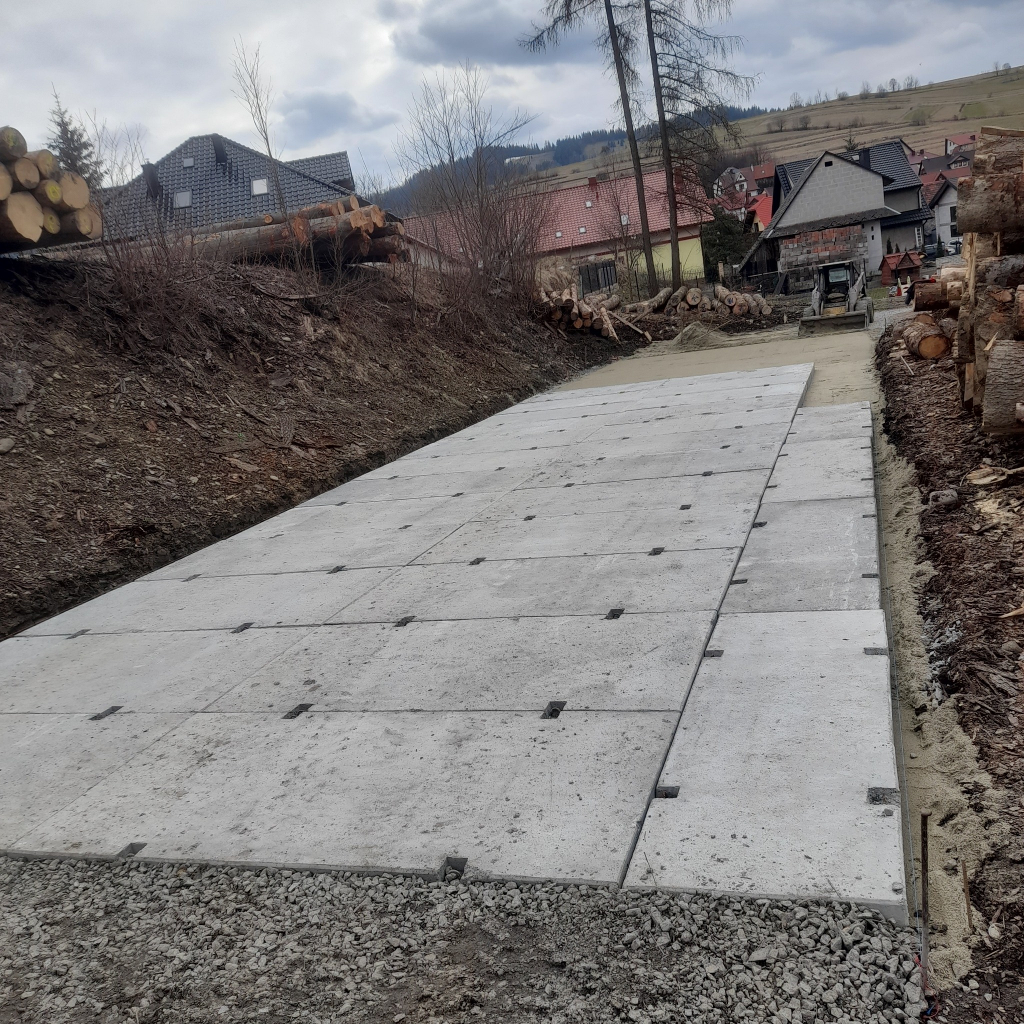 Układanie betonowych płyt drogowych na podsypce żwirowej, widoczny miniładowarka w tle i sterta pni drzew