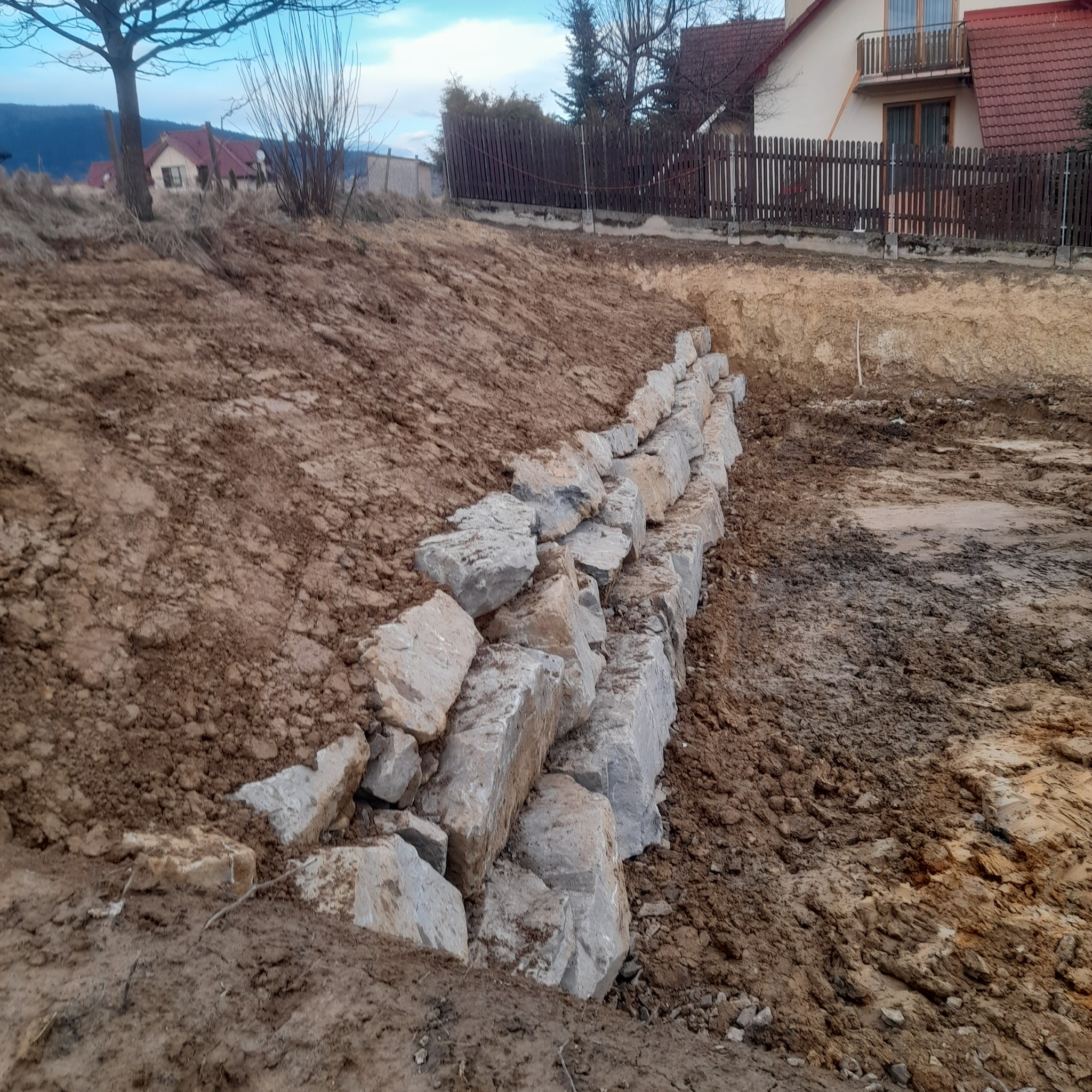 Wznoszenie niskiego muru z szarych kamiennych bloków na nierównym, gliniastym terenie budowy, z widocznymi domami w tle i pochmurnym niebem.