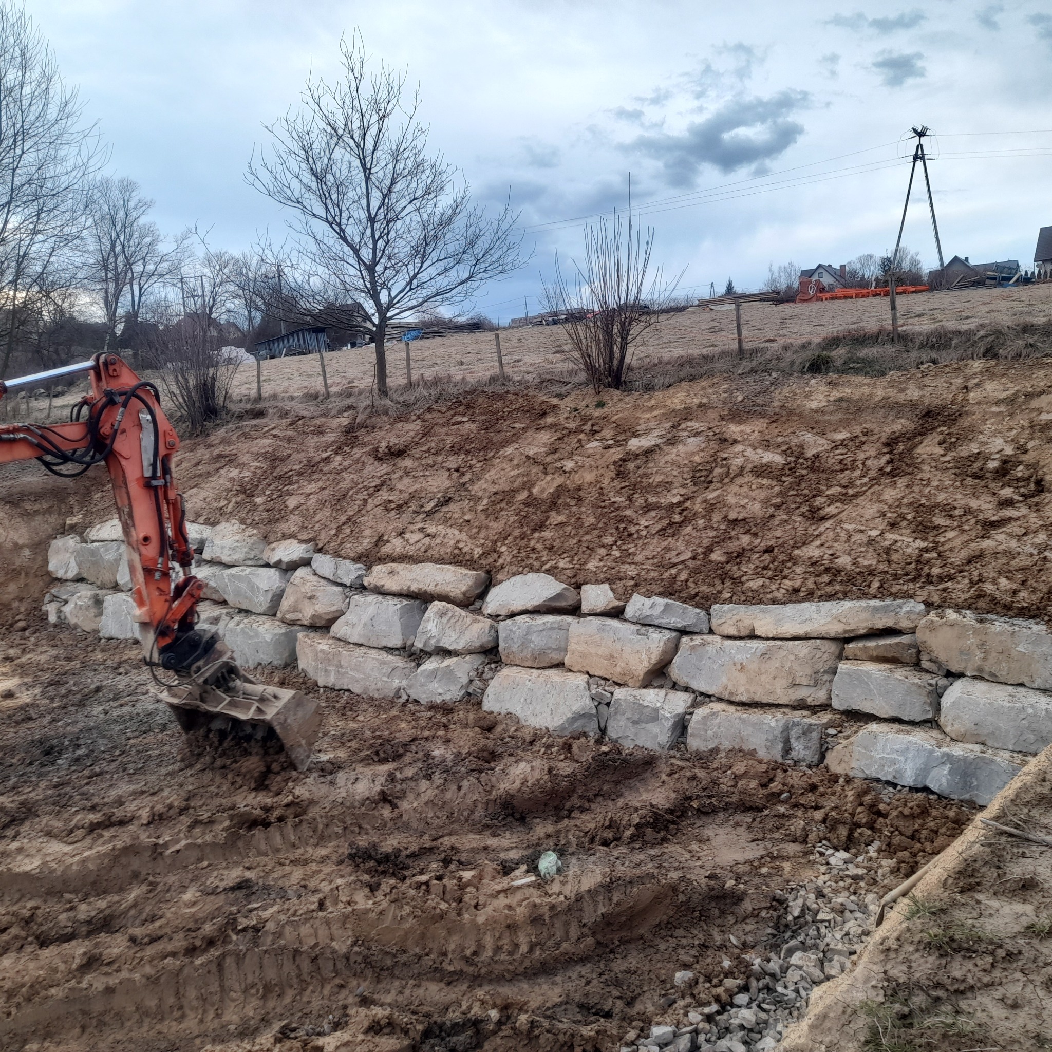 Pomarańczowa koparka układa warstwę z jasnych, nieregularnych kamieni na zboczu, tworząc mur, z widoczną ziemią i gruzem na pierwszym planie oraz drzewami i budynkami w tle pod pochmurnym niebem.